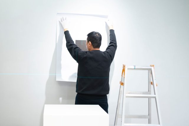 Man placing artwork in Tokyo gallery setting with ladder and gloves.