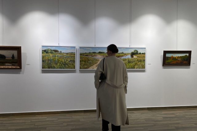 A person in a coat observing landscape paintings in a bright art gallery.